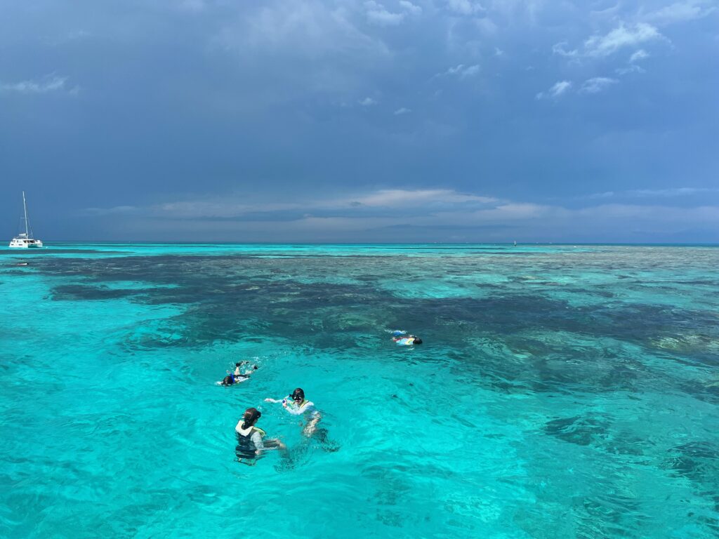 Photograph of Grecian Reef taken from the south end. The water is clear and has snorkelers in the water. You can see a large catamaran boat n the distance